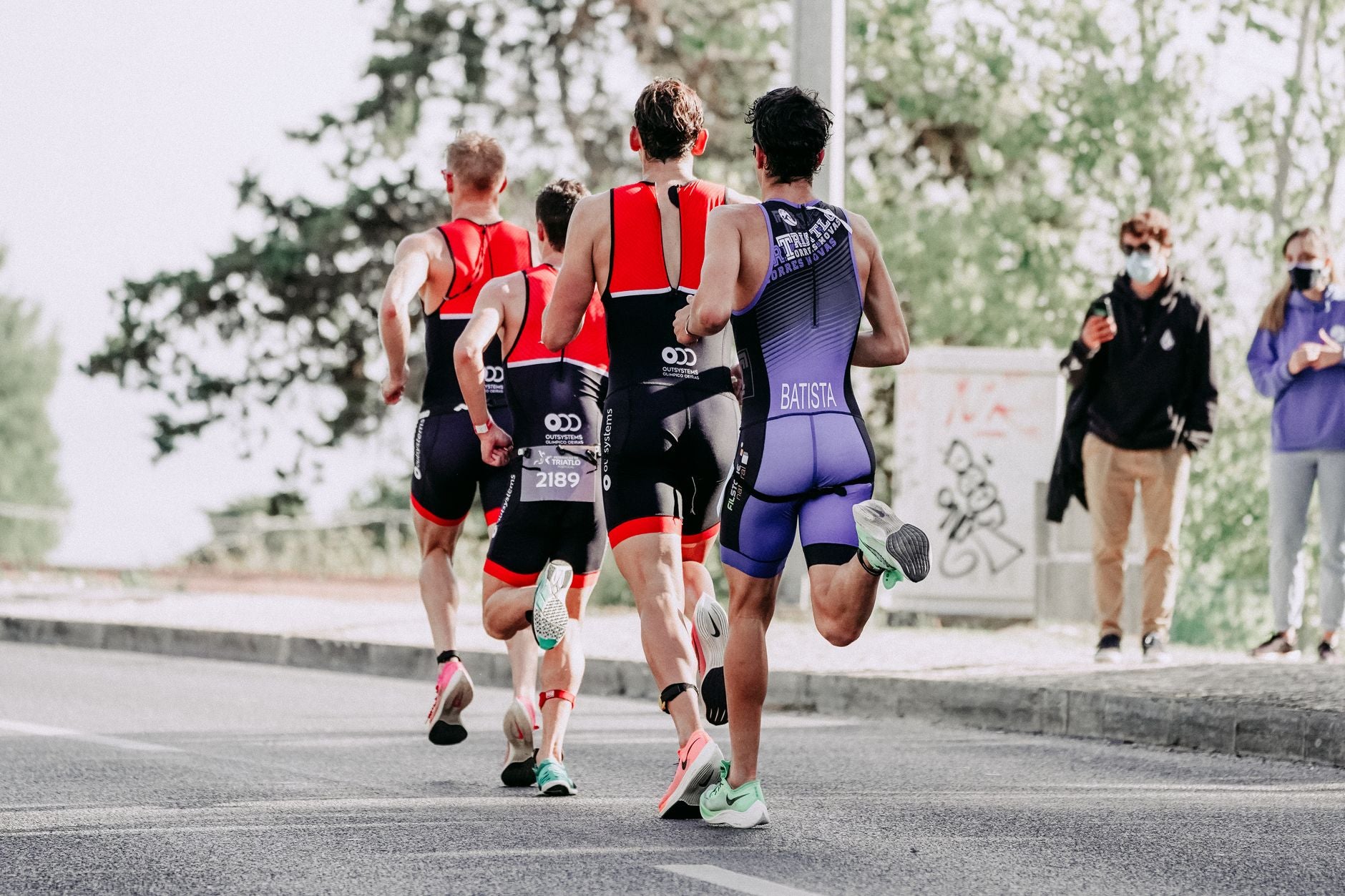 Full body back view of group of sportsmen in track suits running on asphalt road in summertime on blurred background